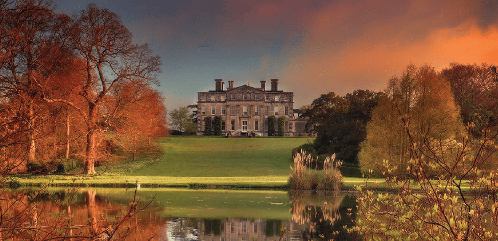 Photo across the lake with the Main House at the top of the lawn, Autumn colours and an orange and pink sky