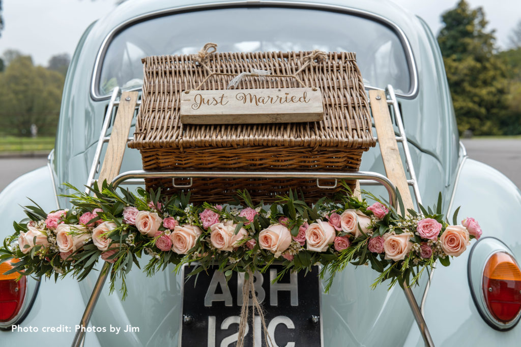 Light blue car with Just Married on a picnic basket on the back, wreath of flowers including light pink roses.
